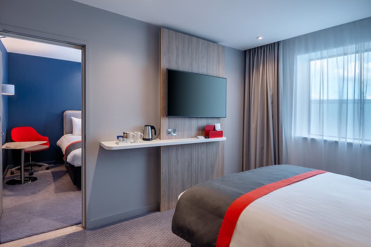 A modern hotel room with a bed, a TV mounted on the wall, a desk with a chair, and a window with curtains.
