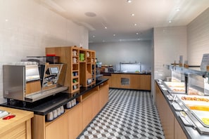 A modern self-service breakfast area with a coffee machine, a variety of mugs, and a buffet with various food items.