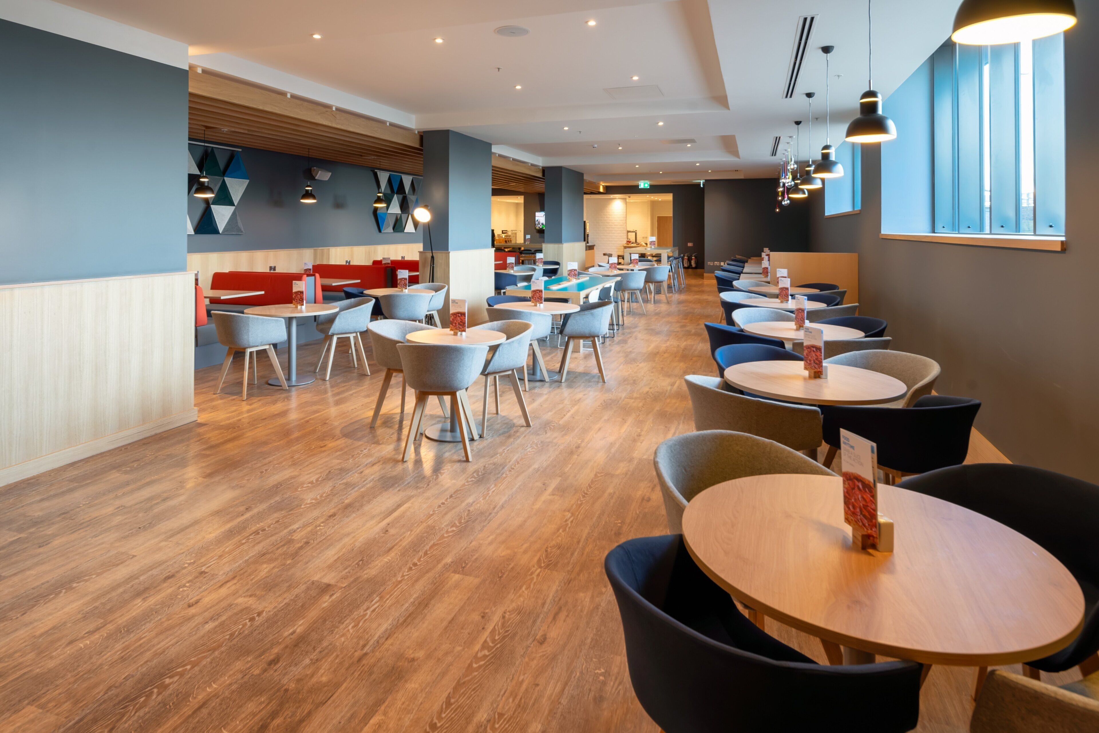 A modern dining area with multiple tables and chairs, wooden flooring, and a mix of red and blue seating.
