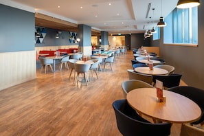 Uma sala de jantar moderna com várias mesas e cadeiras, piso de madeira e assentos em tons de vermelho e azul.