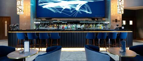 A bar area with a modern design, featuring a long counter, multiple bar stools, and a large screen displaying abstract art.