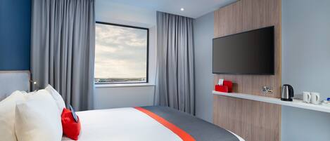 A hotel room with a bed, curtains, a TV, a coffee maker, and a view of the sky.