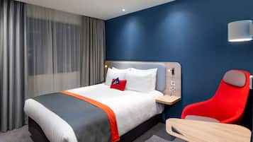 A hotel room with a bed, a red chair, a desk, and a wall-mounted lamp.
