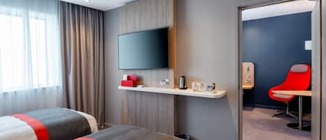 A modern hotel room with two beds, a mounted TV, a small desk with a coffee maker, and a red chair.