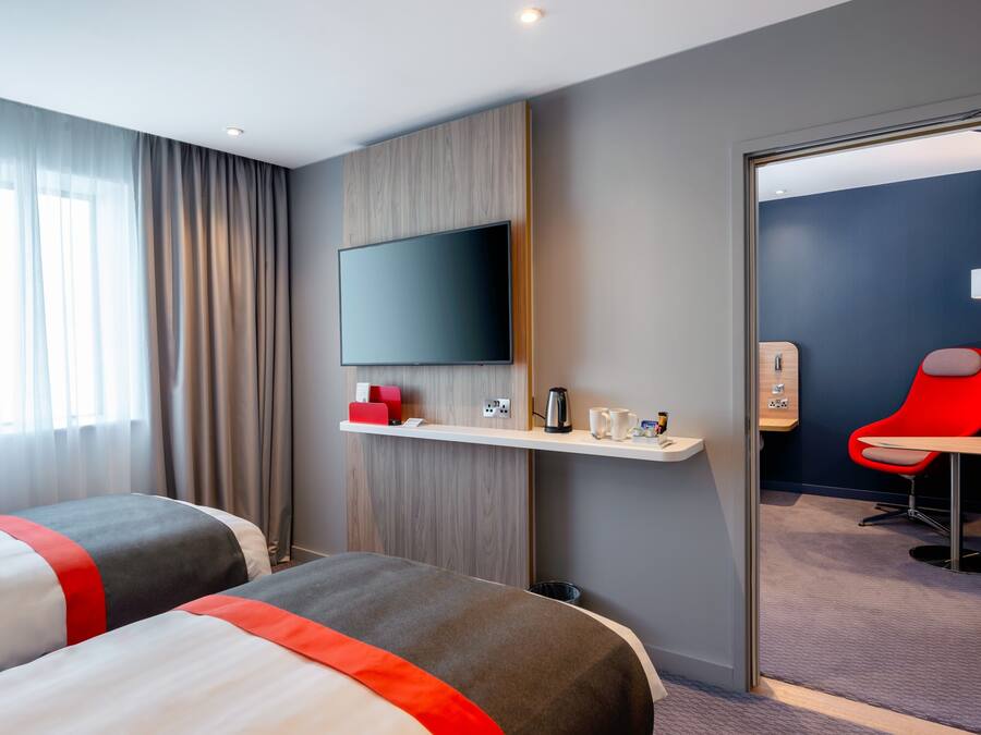 Une chambre d’hôtel moderne avec deux lits, un téléviseur fixé au mur, un petit bureau avec une machine à café et une chaise rouge.