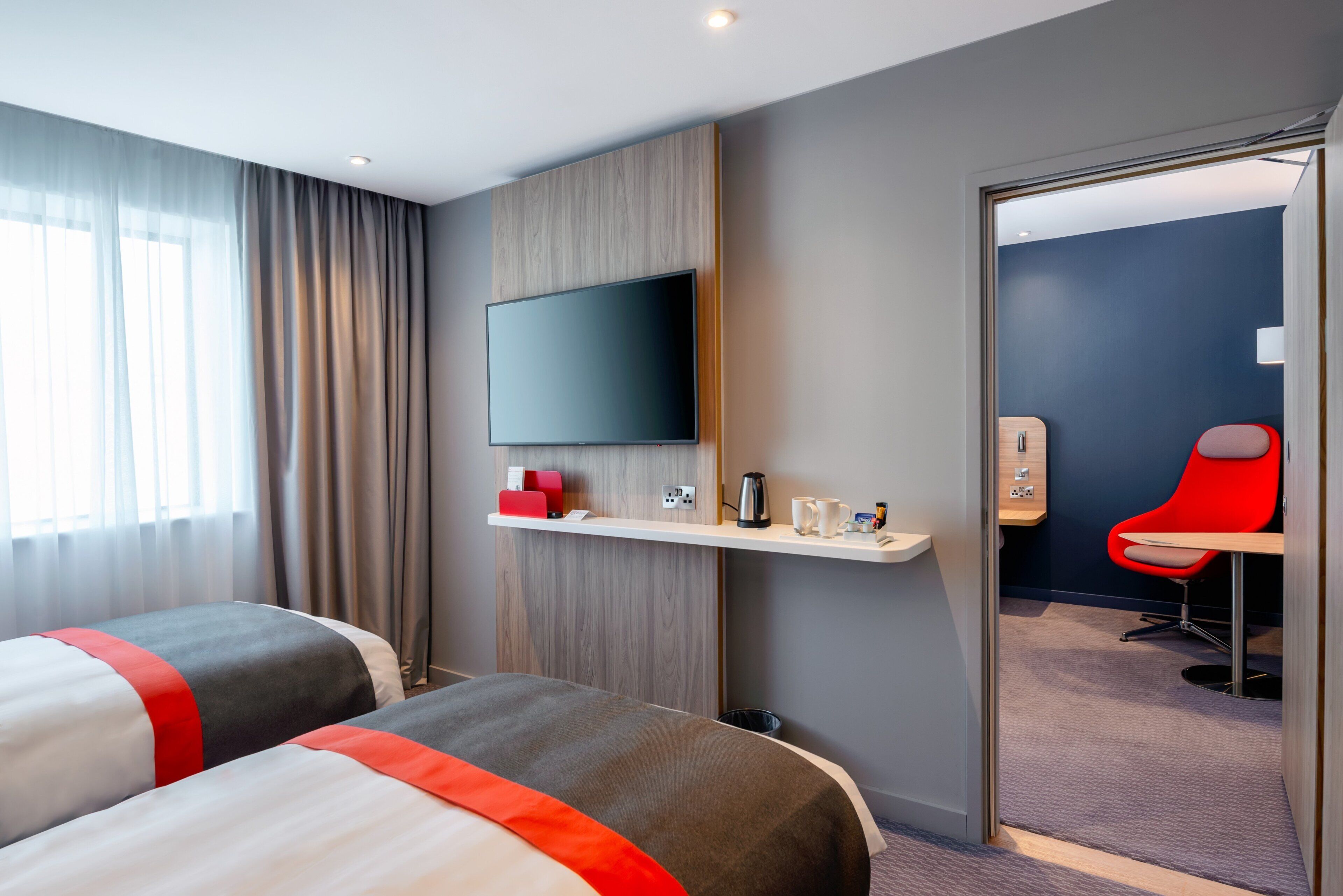 A hotel room with two beds, a TV on the wall, a small desk with a coffee maker, and a red chair.