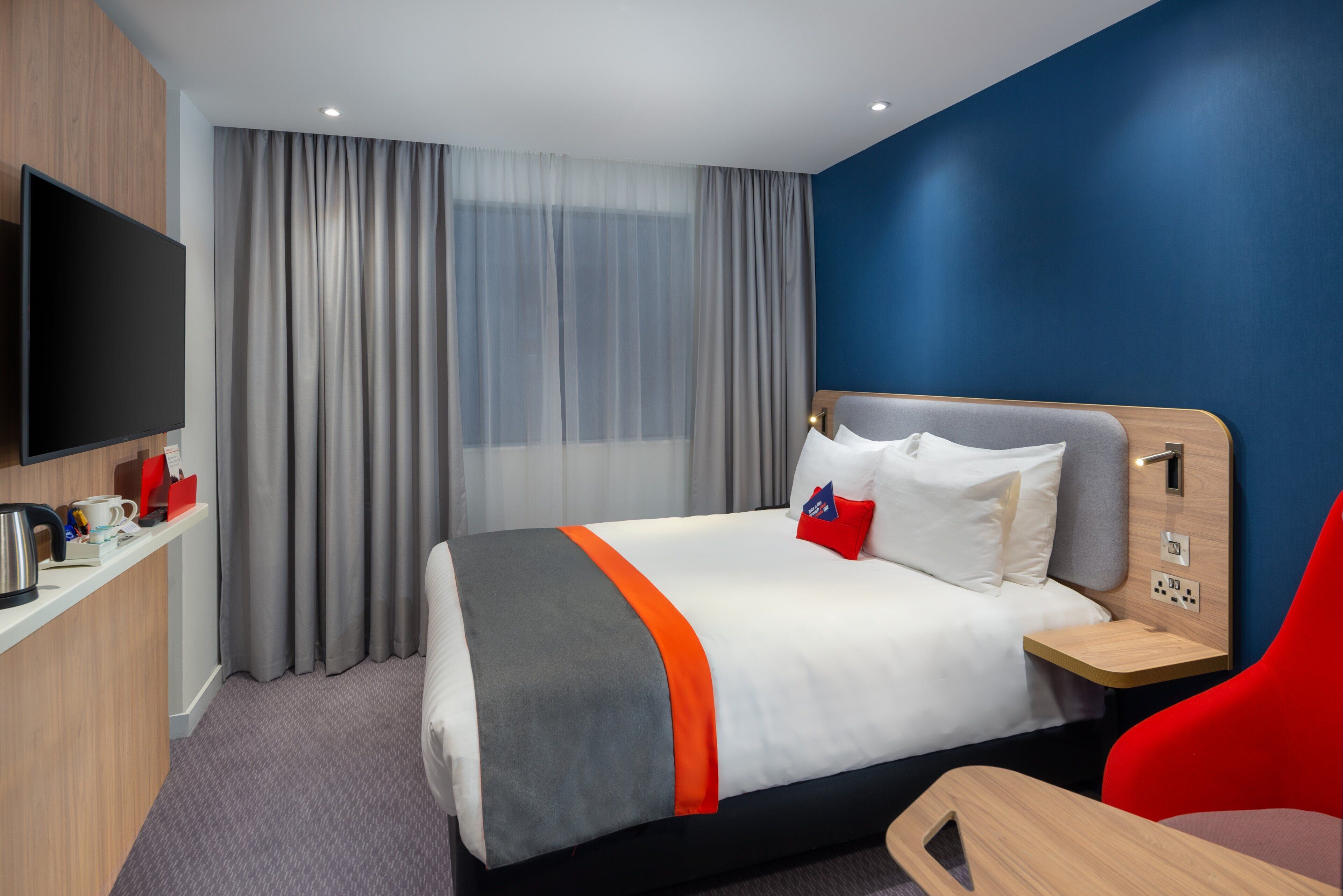 Quarto de hotel com cama, televisão, escrivaninha com cafeteira e uma cadeira vermelha.