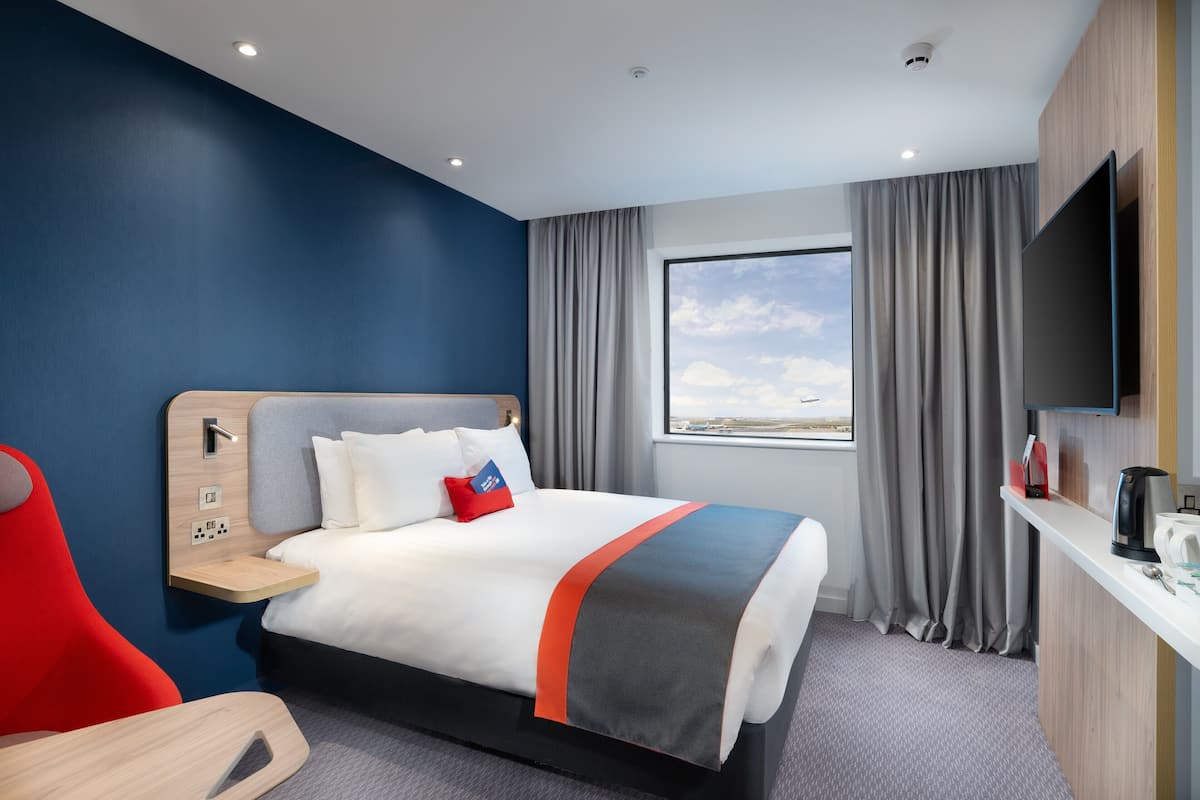 A modern hotel room with a bed, a TV, a desk, and a window with curtains.