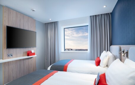 A hotel room with two beds, a flat-screen TV mounted on a wooden panel, a window with curtains, and a view of the landscape.