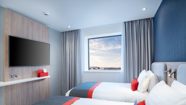 A hotel room with two beds, a flat-screen TV mounted on a wooden panel, a window with curtains, and a view of the landscape.