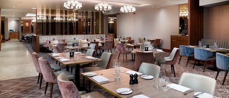 A modern dining area with multiple tables and chairs, chandeliers, and a patterned carpet.