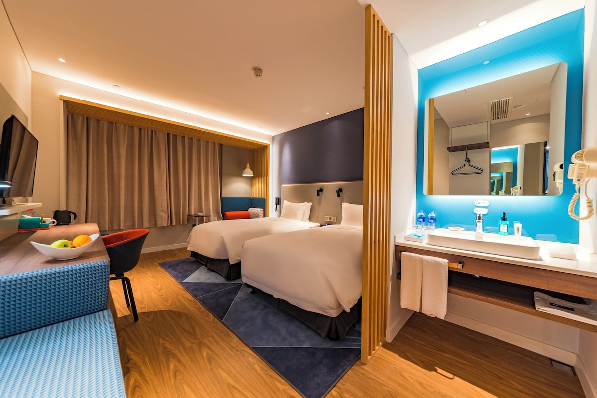 A modern hotel room with two beds, a sitting area with a bowl of fruit, a large mirror, and a bathroom with a sink and toiletries.