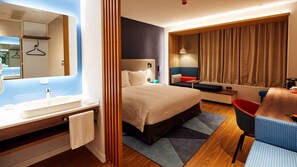 A modern hotel room with a large bed, a desk, a chair, a bathroom with a sink and mirror, and a view of the city.