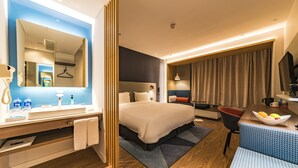 Een moderne hotelkamer met een groot bed, een zithoek met een blauwe sofa met een patroon, een houten vloer, en een badkamer met een grote spiegel en een wastafel.