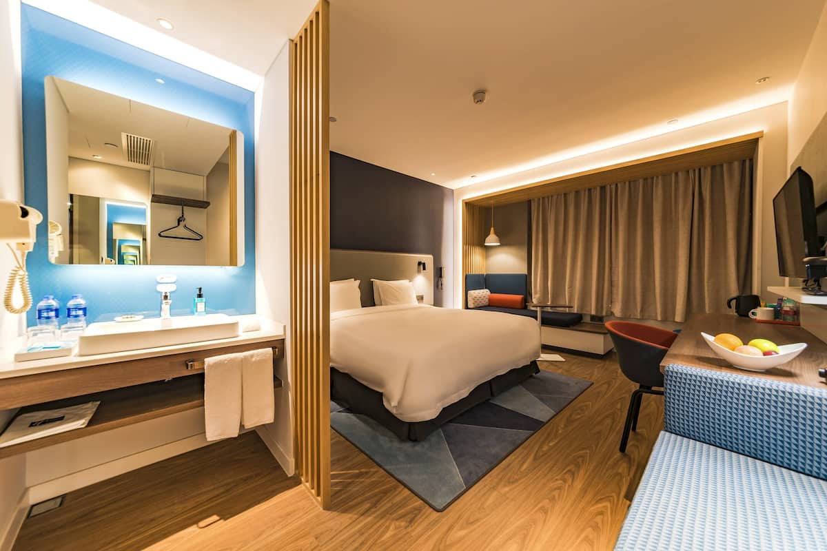 Une chambre d’hôtel moderne avec un grand lit, un coin salon doté d’un canapé bleu à motifs, un plancher en bois et une salle de bain avec un grand miroir et un lavabo.