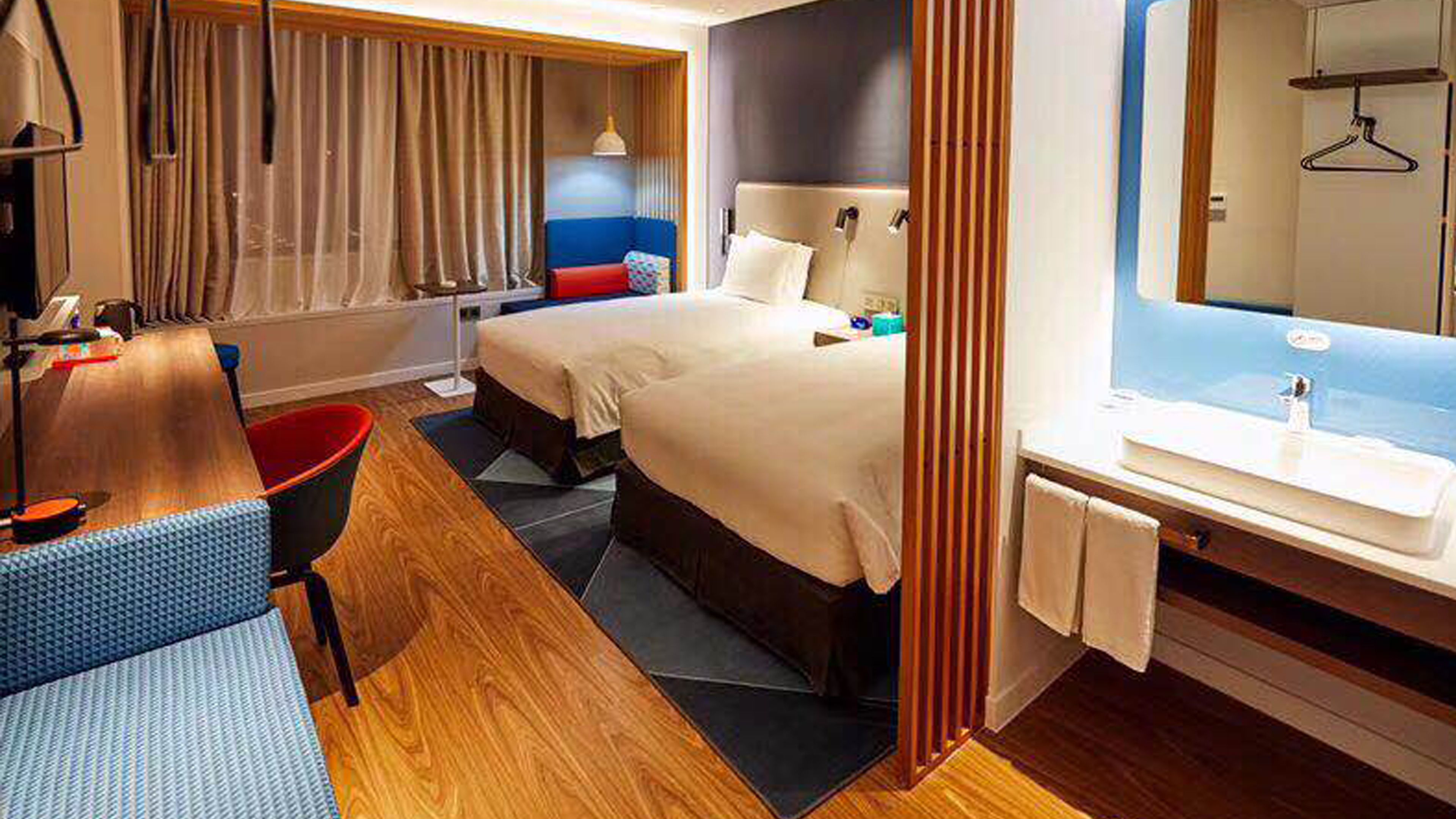 Habitación de hotel con dos camas, un escritorio, una silla, baño con espejo y lavamanos, y ventana con cortinas.