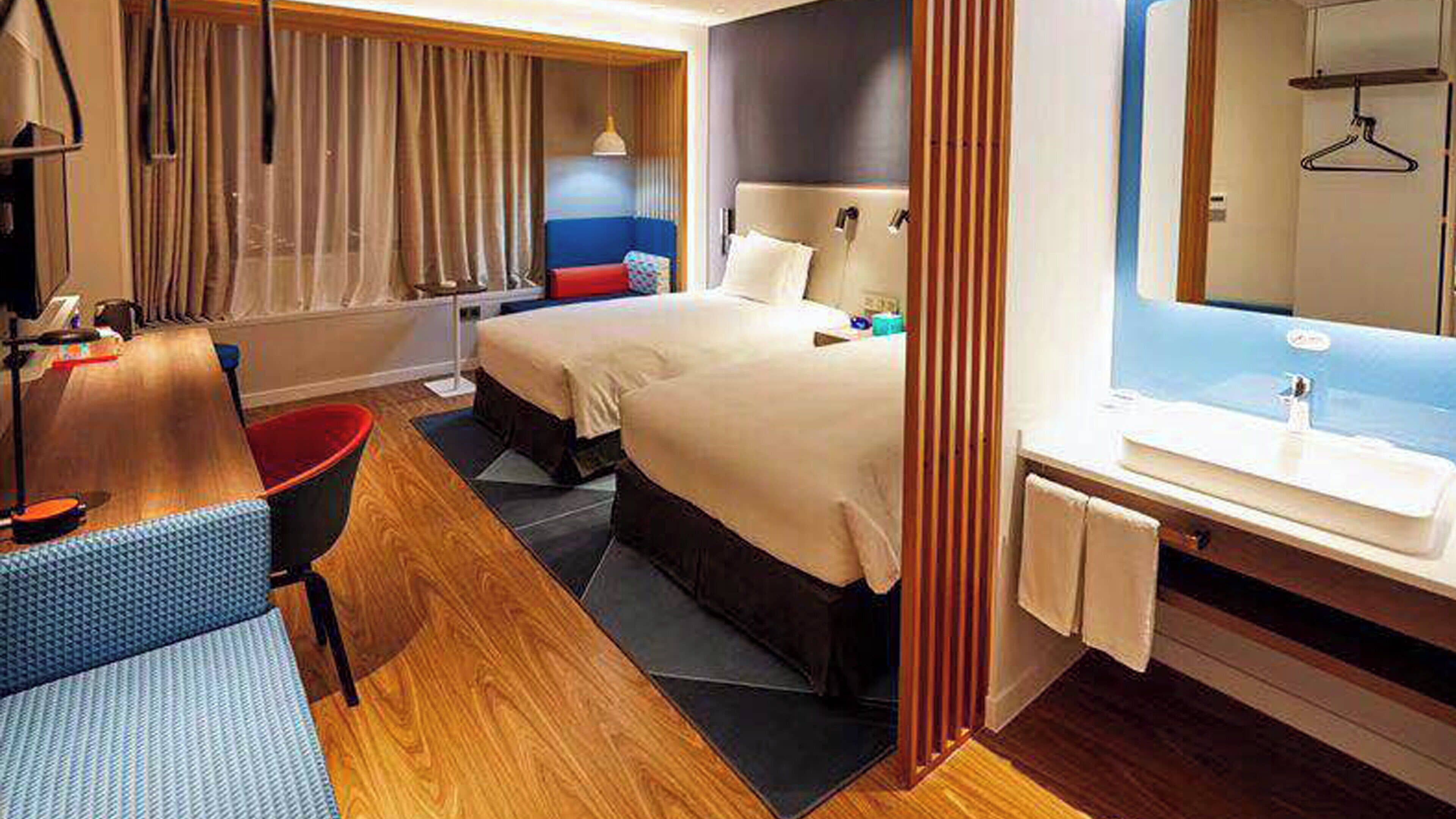 Habitación de hotel con dos camas, un escritorio, una silla, un baño con espejo y una ventana con cortinas.