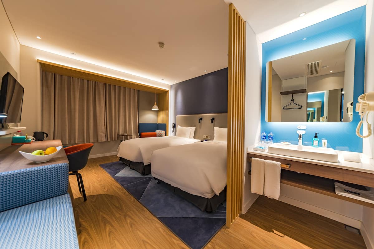 Une chambre d’hôtel moderne avec deux lits, un coin salon agrémenté d’un panier de fruits, un grand miroir et une salle de bain équipée d’un lavabo et d’articles de toilette.