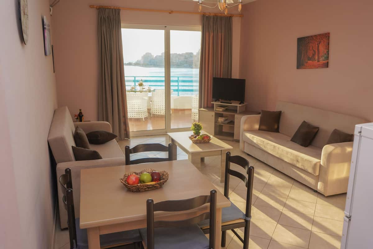 A living room with a sofa, a dining table with chairs, a TV, and a view of the sea through a large window.