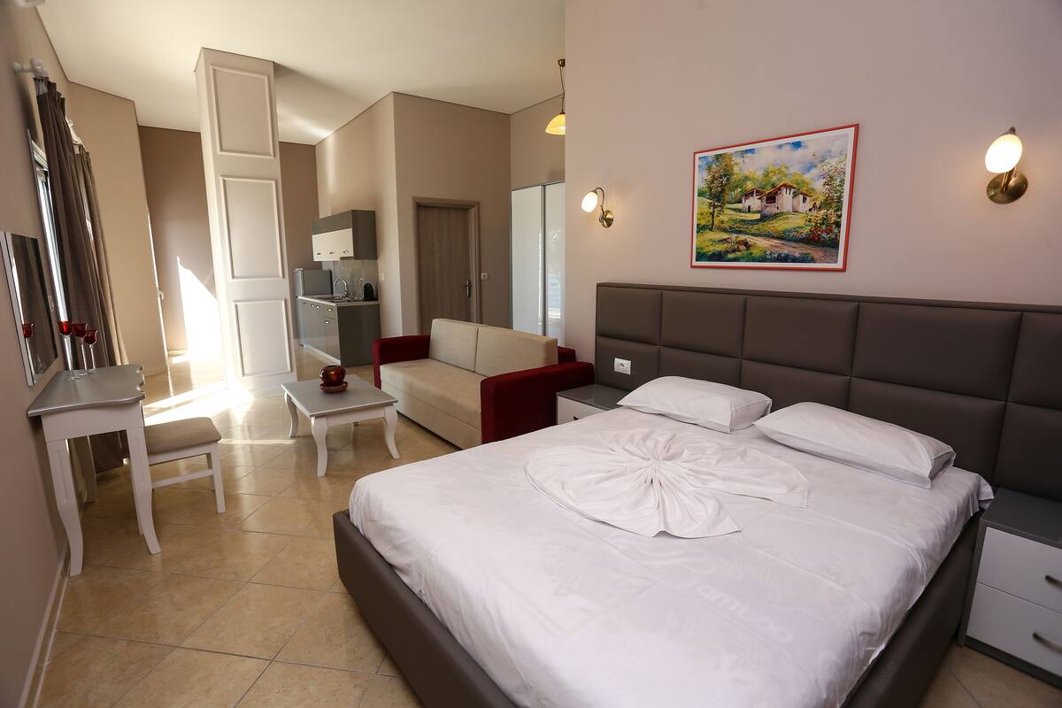 A hotel room with a bed, bedside tables, a desk, a chair, a sofa, a wardrobe, and a painting on the wall.