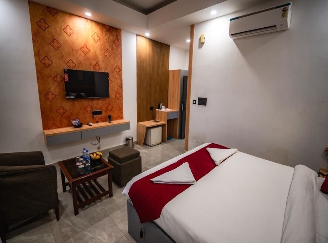 A hotel room with a bed, a TV mounted on the wall, a small table with a bottle, a chair, and a wooden cabinet.