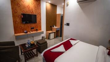 A hotel room with a bed, a TV mounted on the wall, a small table with a bottle, a chair, and a wooden cabinet.