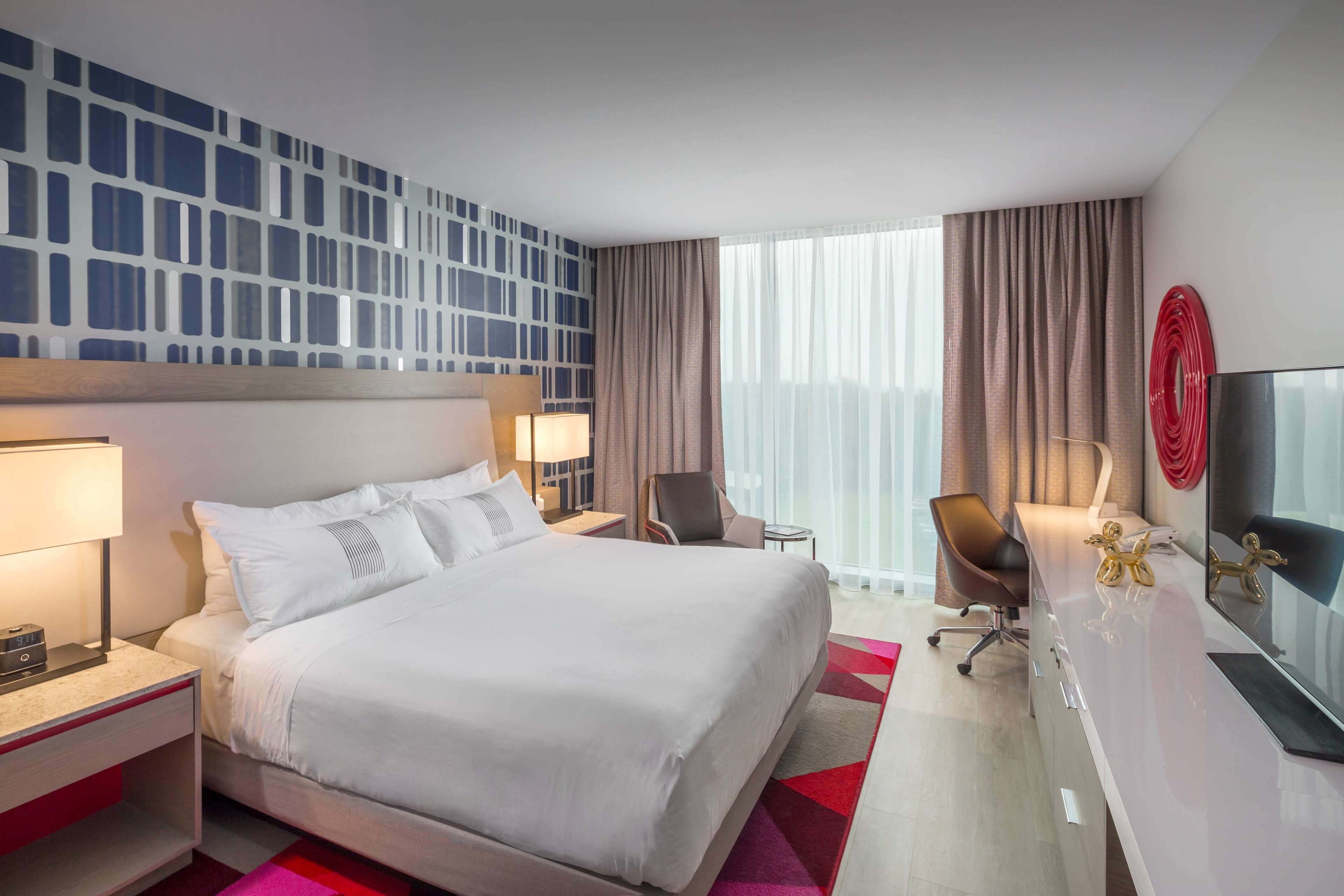 A hotel room with a large bed, two bedside tables with lamps, a desk with a TV, a chair, and a large window with curtains.