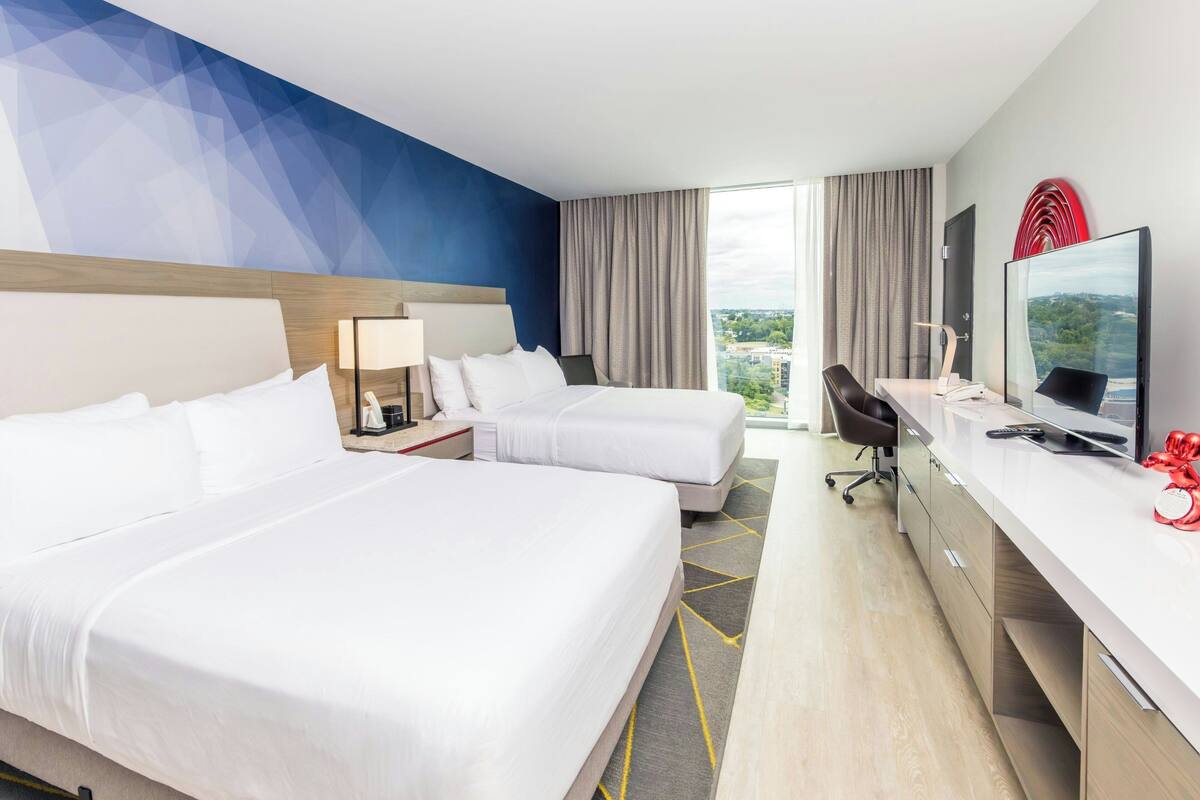 Habitación de hotel con dos camas, un escritorio, una silla, un televisor y un ventanal con vistas.