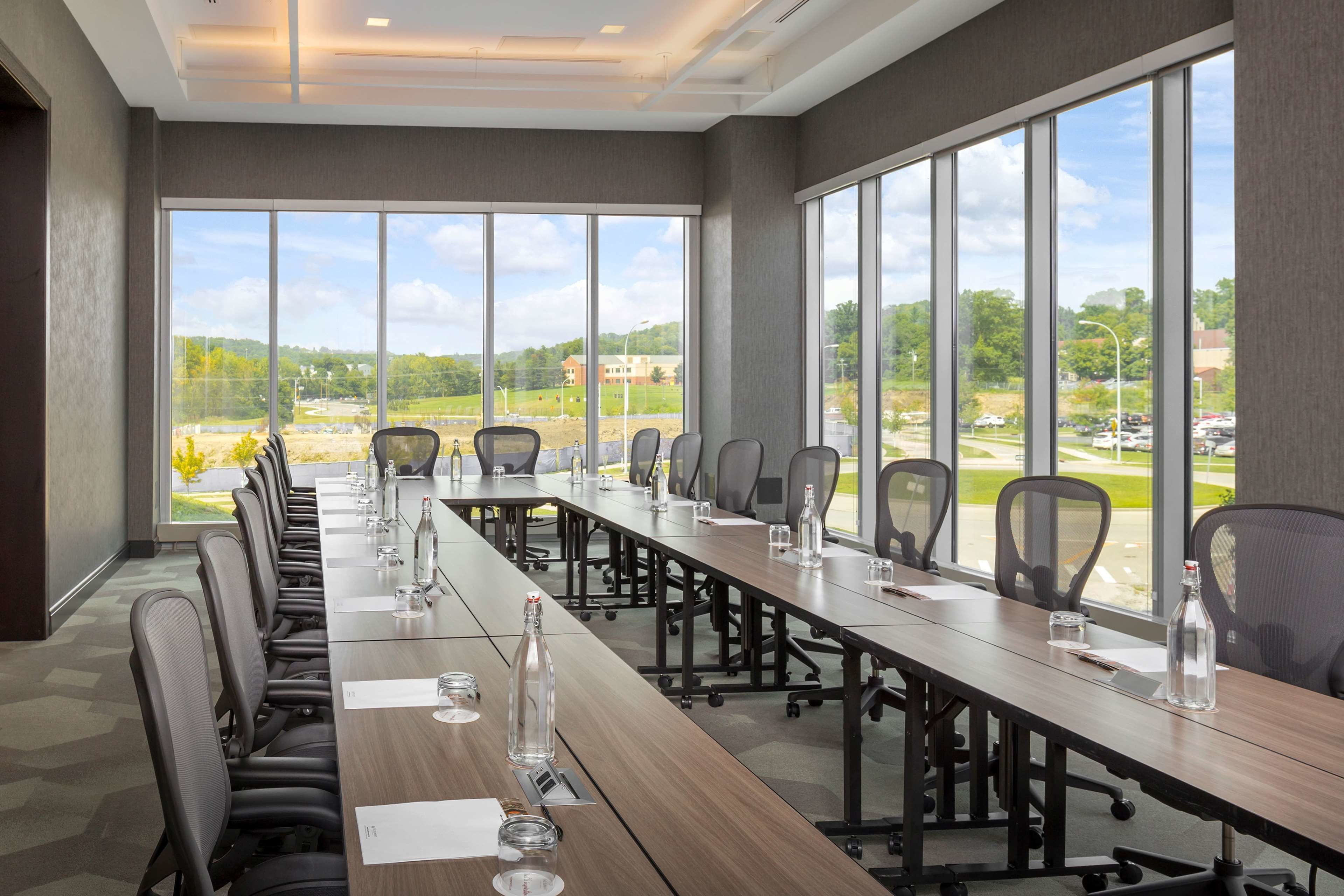 A conference room with a long table, chairs, and large windows offering a view of a parking lot and buildings.
