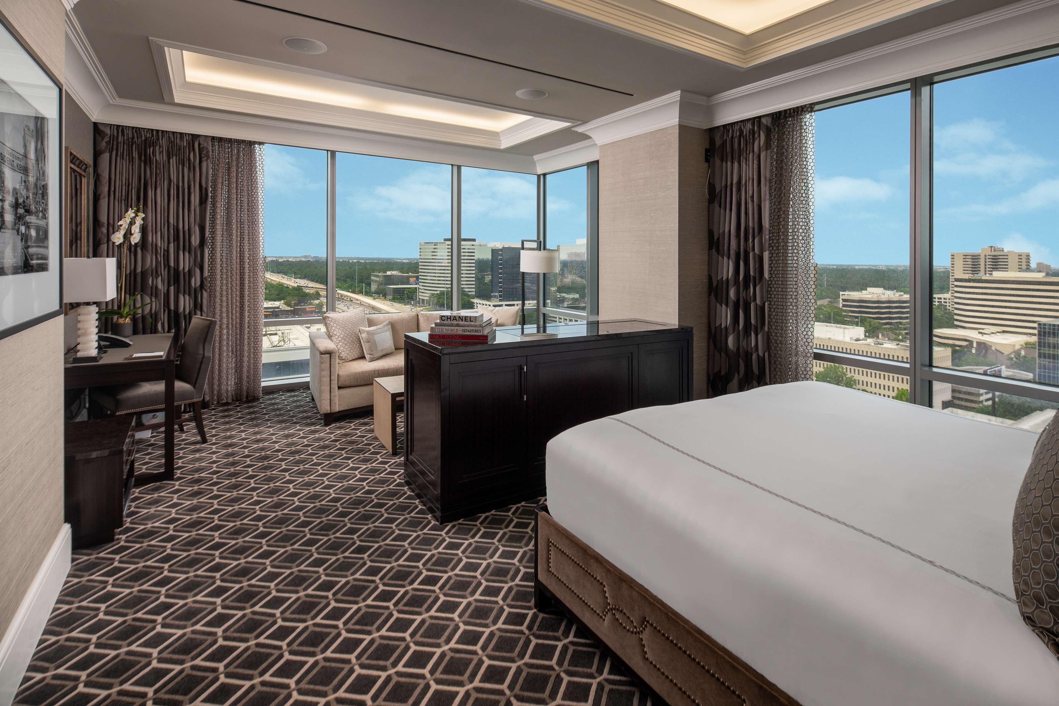 A hotel room with a large bed, a desk, a chair, and a sofa. There are large windows with a city view.