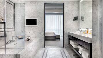 Un baño moderno con un espejo grande, una bañera, un lavabo y una ducha. Hay un televisor montado en la pared y una ventana con persianas.