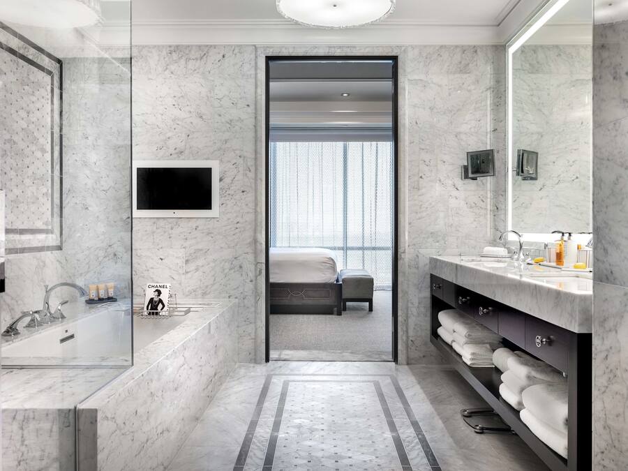 A modern bathroom with a large mirror, a bathtub, a sink, and a shower. There is a television mounted on the wall and a window with blinds.