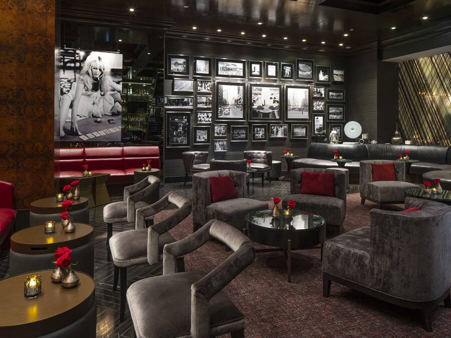 A modern lounge area with leather sofas, armchairs, and round tables, featuring a wall of black-and-white photographs and a large TV screen.