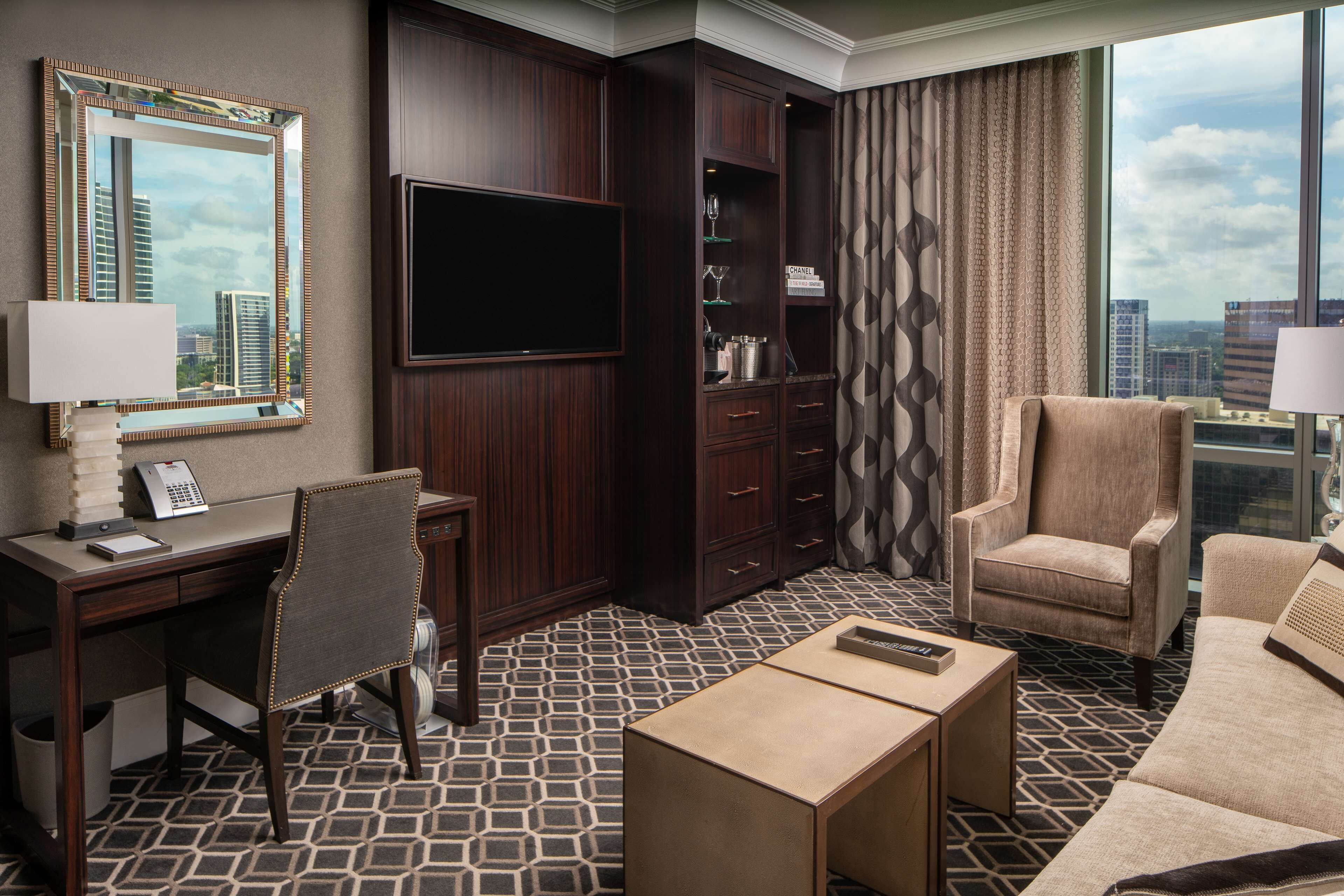 A hotel room with a large TV, a desk with a telephone, a chair, a sofa, and a patterned floor.