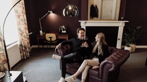A living room with a dark-colored sofa, a coffee table, a lamp, and a fireplace. - Châteauhotel en restaurant De Havixhorst (De Schiphorst)