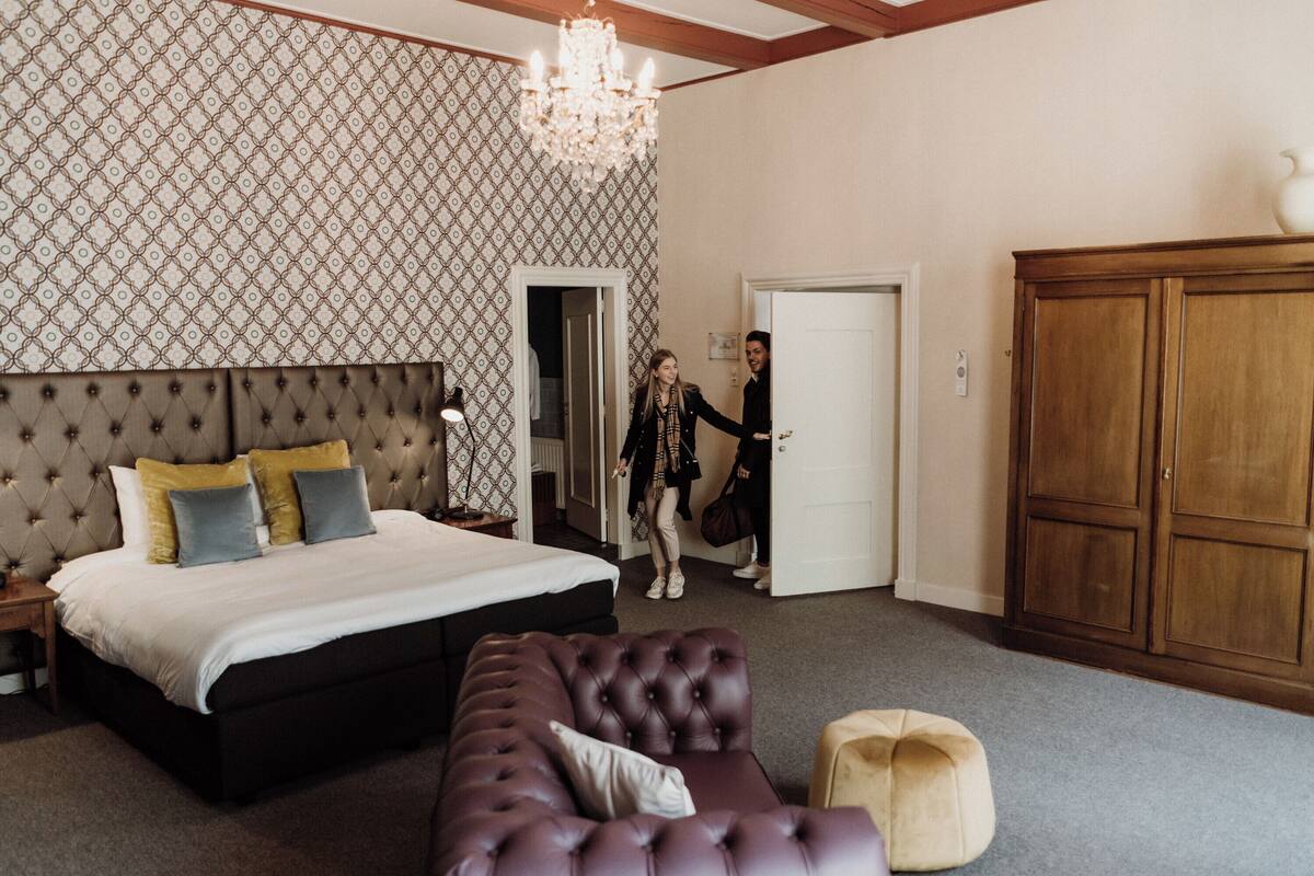 Quarto com uma cama grande, cabeceira estofada, lustre, guarda-roupa e uma porta que dá para outro cômodo.