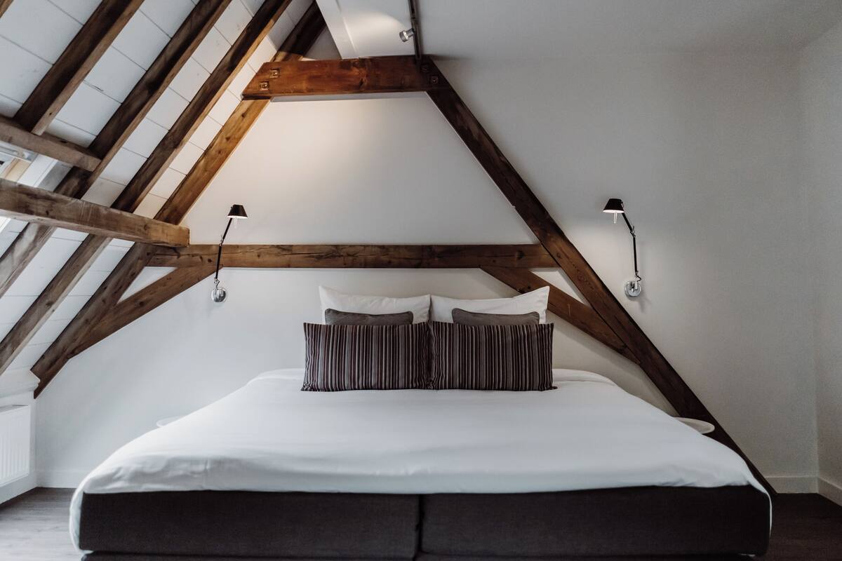 Quarto com teto inclinado, vigas de madeira, cama grande com almofadas brancas e listradas, e duas luminárias fixadas na parede.