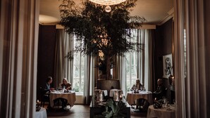 A restaurant with round tables, a large tree centerpiece, and a chandelier. - Châteauhotel en restaurant De Havixhorst (De Schiphorst)