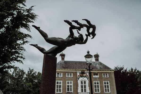 Una estatua de una figura humana en una pose dinámica, frente a un edificio histórico con múltiples ventanas y un farol tradicional.