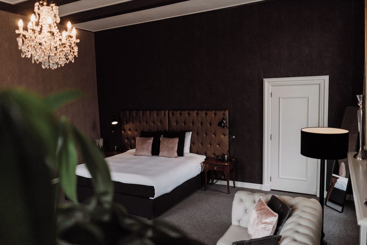 Quarto com cama grande, cabeceira estofada, mesa de cabeceira, abajur, porta e sofá.