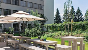Un patio al aire libre con mesas y bancos de madera, rodeado de vegetación y un edificio al fondo.