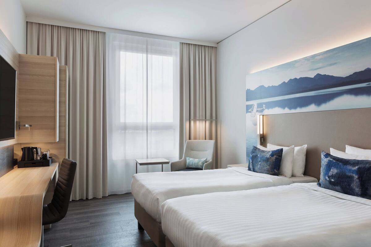Une chambre d’hôtel avec deux lits, un bureau, une chaise, une télévision et un tableau représentant des montagnes.