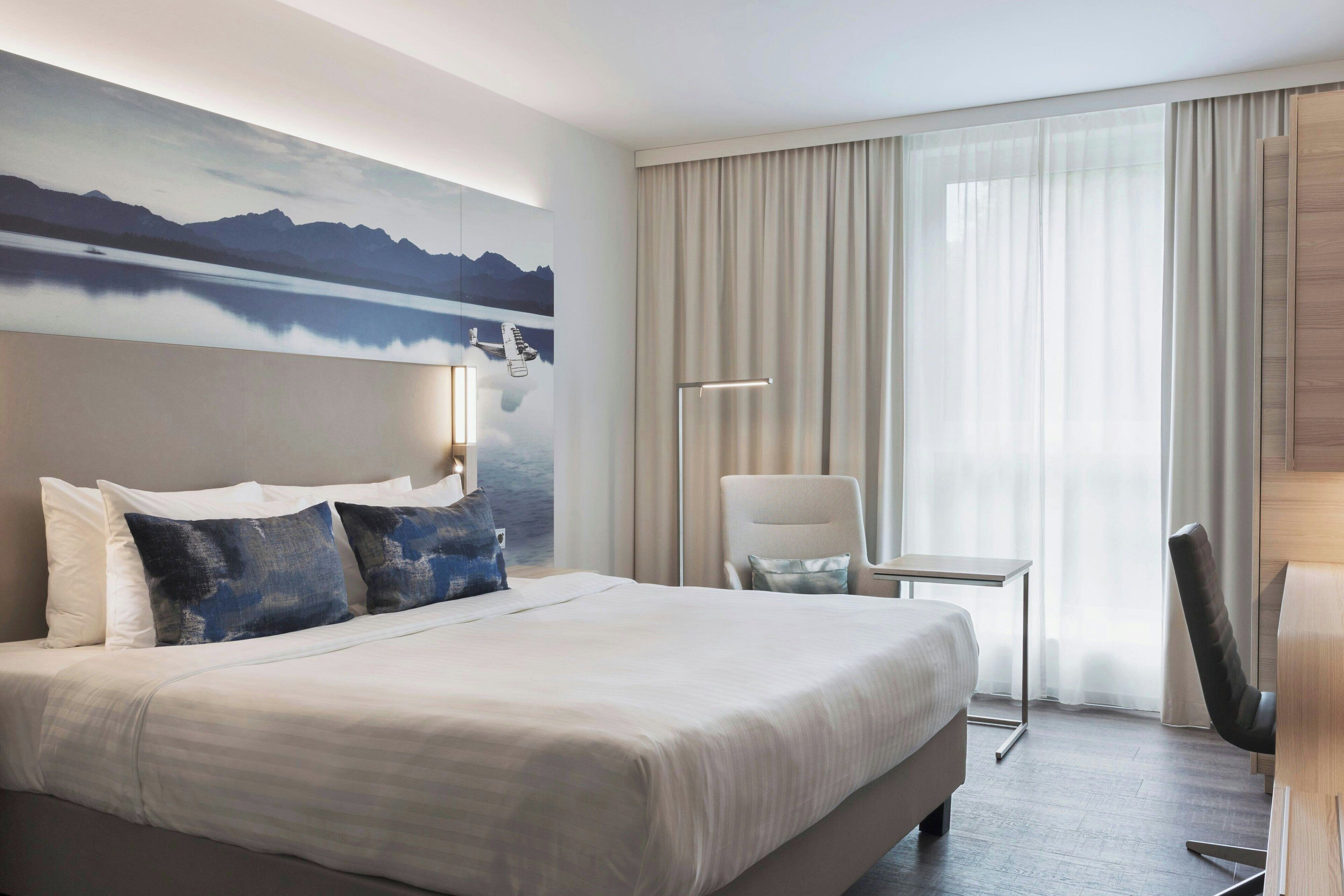A hotel room with a large bed, a desk, a chair, a painting of mountains and a lake, and a view of the outdoors through sheer curtains.