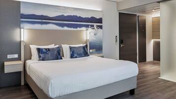 Una habitación de hotel moderna con una cama grande, una mesita de noche, una lámpara de pared y una puerta a la derecha.