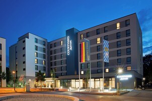 Um hotel Courtyard Marriott com design moderno, com vários andares e uma entrada destacada com letreiros iluminados.