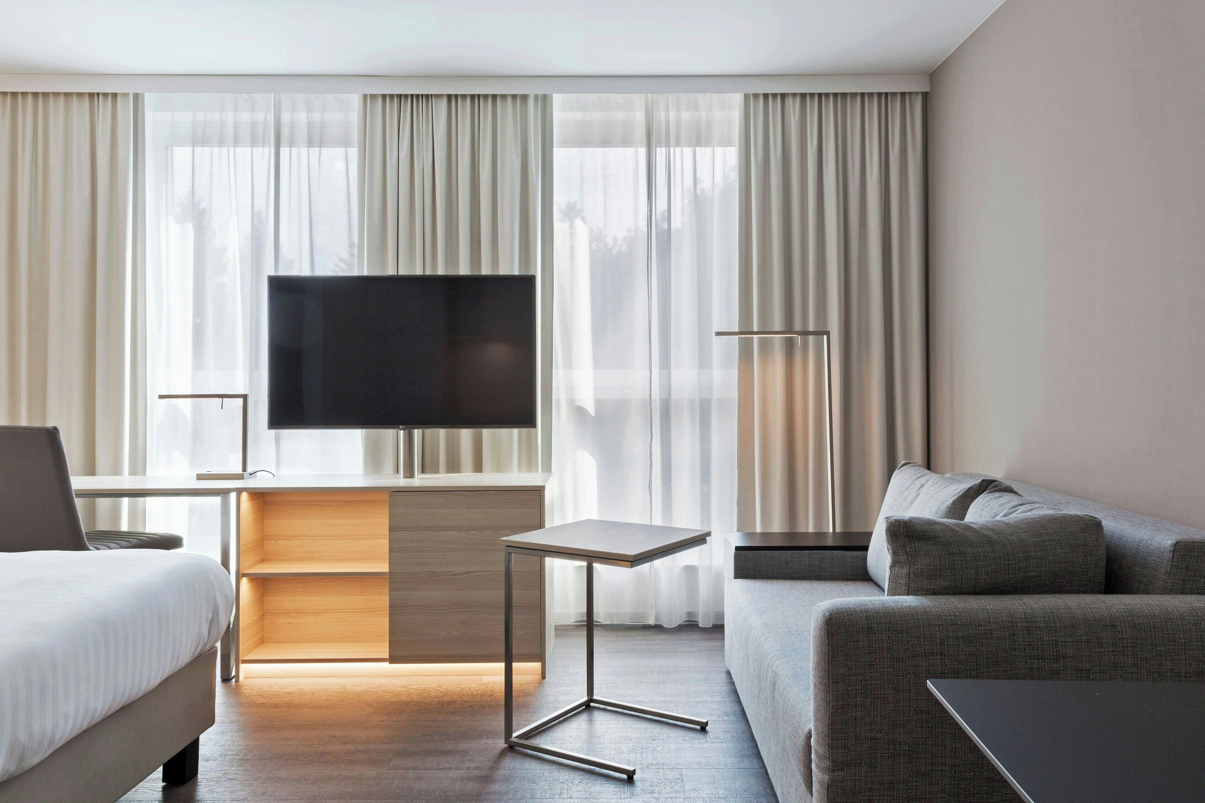 Moderni hotellihuone, jossa on sänky, sohva, televisio, työpöytä ja lamppu.