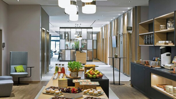 Un moderno vestíbulo de hotel con un largo mostrador de recepción, un área de descanso con una silla gris y un bufé con diversas opciones de comida.