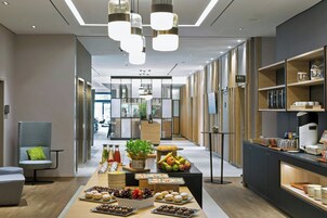 Um lobby moderno de hotel com um balcão de recepção alongado, uma área de estar com uma cadeira cinzenta e um buffet com diversas opções de comida.