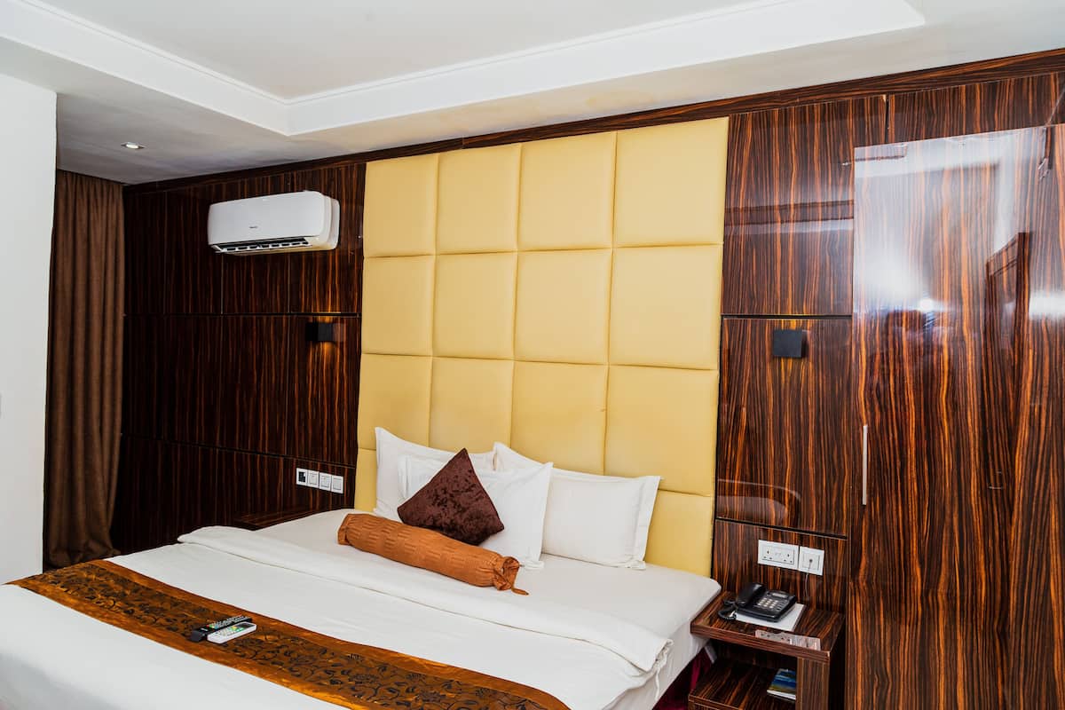 Ein Hotelzimmer mit einem großen Bett, einem Holzkopfende, einer Klimaanlage, einem Nachttisch mit Telefon und einer an der Wand montierten Steckdose.