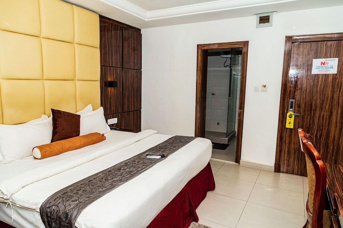 Ein Hotelzimmer mit einem großen Bett, einem Holzschrank, einer Tür mit einem Rauchverbotsschild und einem durch eine offene Tür einsehbaren Badezimmer.